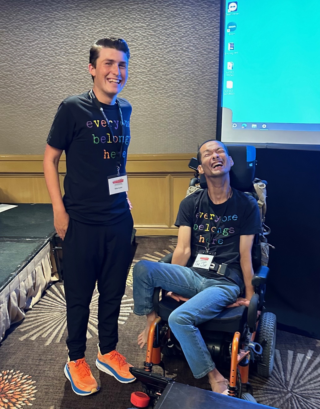two men wearing short sleeve black t-shirts with the words "Everyone Belongs Here" printed in multicolored font. The young caucasian man named Otto is on the left and smiling broadly with Tim next to him. Tim is an Asian man also smiling broadly seated in a motorized wheelchair. Both men have short brunette hair stylishly gelled back. They are wearing lanyards from the conference they are presenting at together. This is a picture of great joy.
