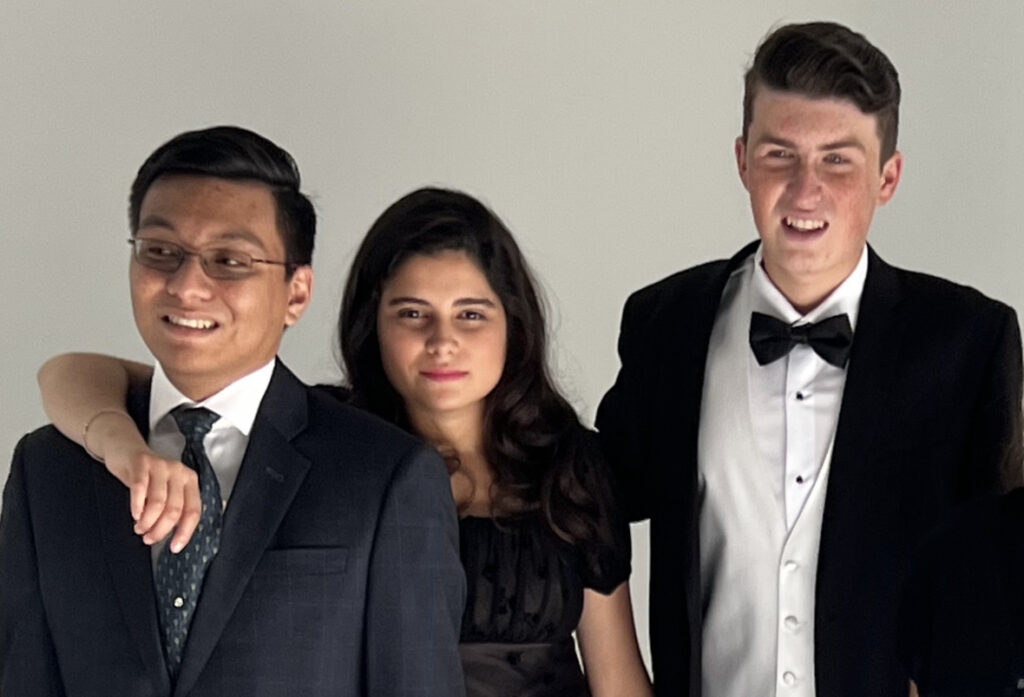 Three young adults standing in front of a white backdrop. William, a young Latin/Filipino man , on the far left, is wearing wire rimmed glasses, has short black hair gelled back and a charcoal suit, white shirt and navy tie with a small pattern, and he is smiling. Bella, the young Latin woman in the center has her left arm draped on William's shoulder. She is wearing a black short sleeved dress, has shoulder length brunette hair and she too is smiling wearing pink lipstick. Otto, on the right is a young caucasian man with a broad toothy grin, wearing a black tuxedo, white shirt, white vest with a black bowtie. His short brunette hair is gelled back.