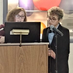Corissa co-presenting with State Lead, Mary Shehan. Both are standing at a podium and speaking into a microphone. Corissa is wearing a black suit top and khaki pants.
