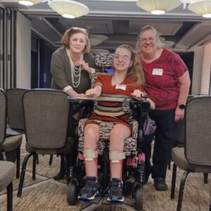 Jeanie with her mom and state lead, Mary. Jeanie is a wheelchair and her mom and Mary are standing on each side of her.