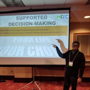 Kaiden Tolbert presenting at the Michigan statewide Council on Exceptional Children Conference in 2022. He is standing in front of a screen and pointing to it. The words on the screen say: Supported Decision Making